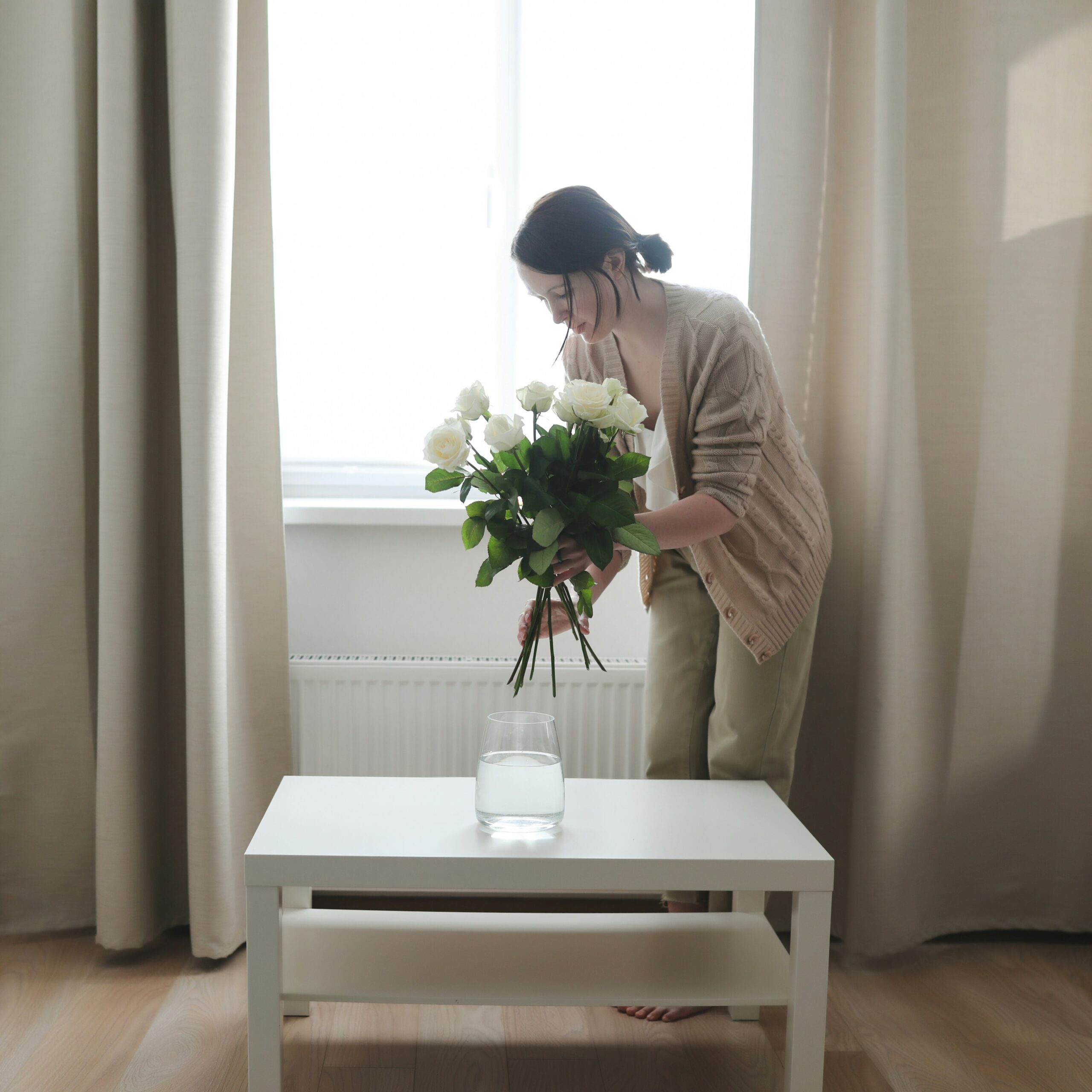 A woman arranging a bouquet of white roses into a glass vase filled with water on a white coffee table. She is in a softly lit room with beige curtains, standing near a window with natural light streaming in